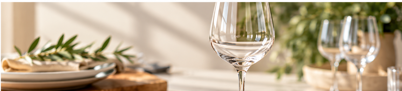 A clear wine glass on a warmly lit table set with plates, linen and greenery