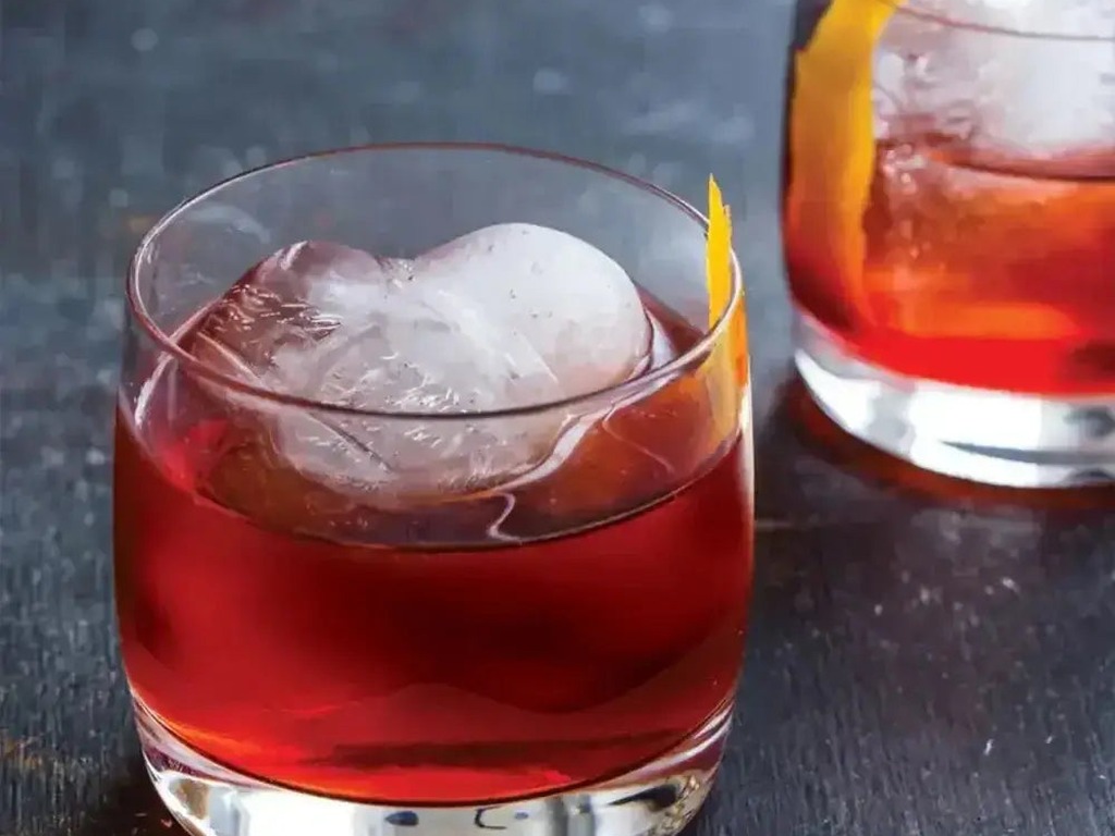 Zoku oversized ice spheres in two red cocktails with citrus twists on a dark bar top