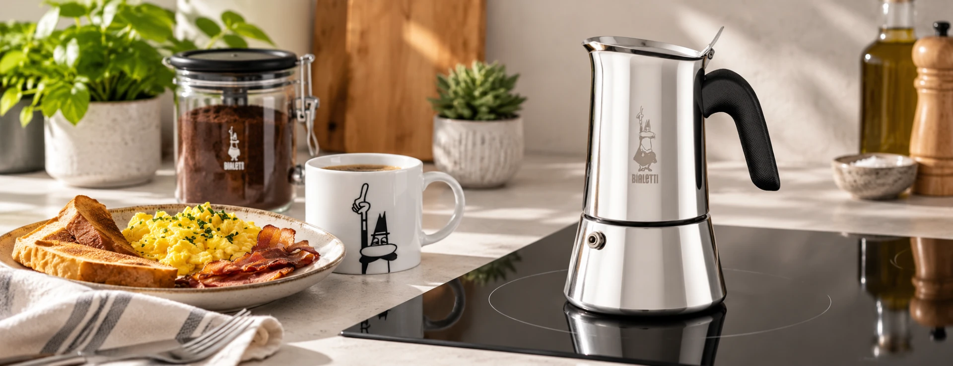 Stainless-steel Bialetti moka pot on a black induction hob in a warm kitchen with coffee and breakfast nearby.