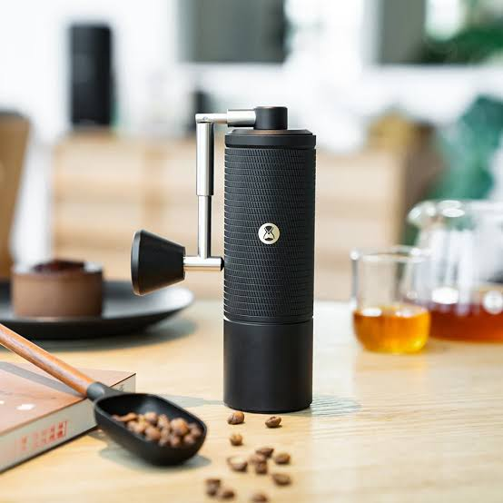 A premium black manual burr coffee grinder on a wooden table with scattered coffee beans