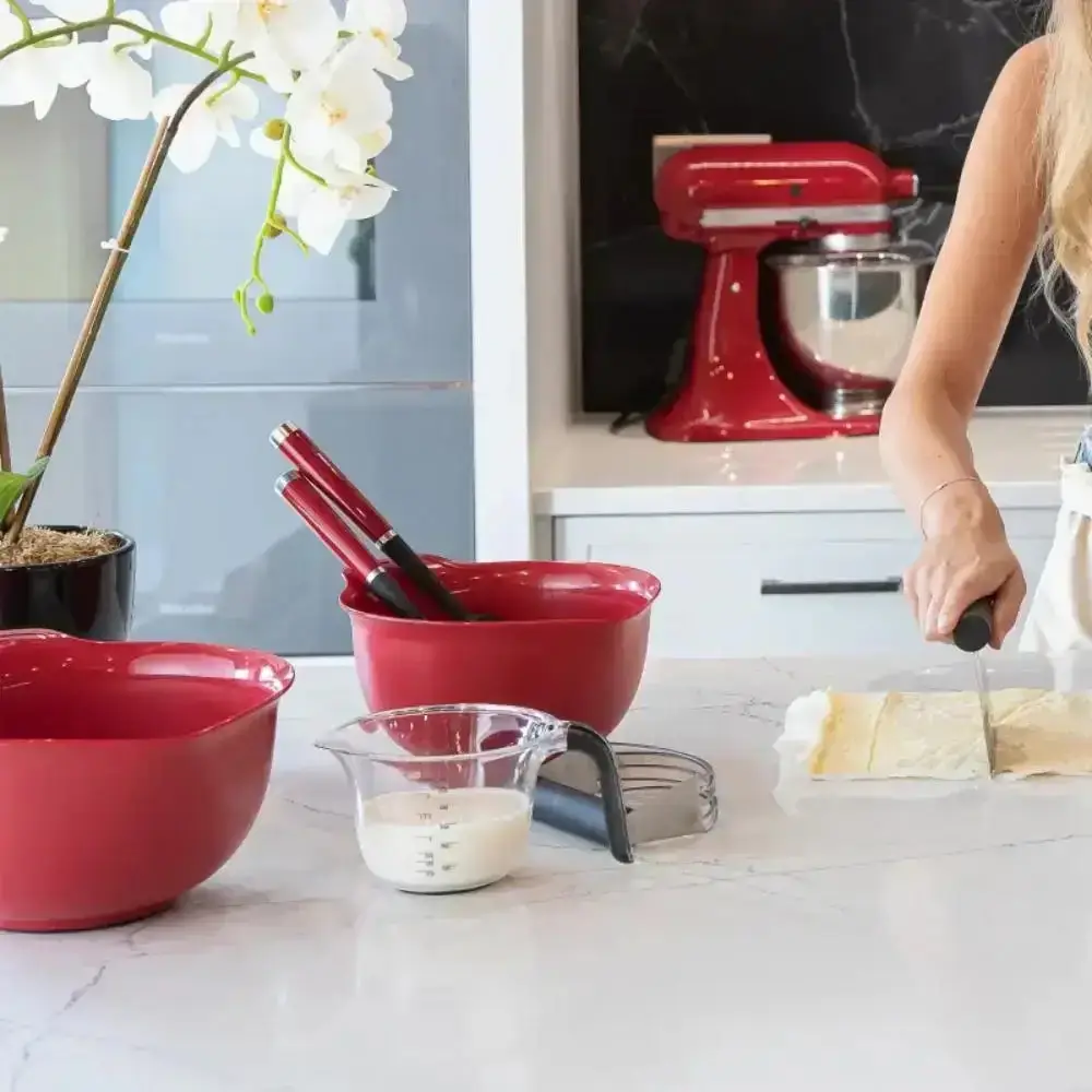 Red mixing bowls, a measuring jug, whisk and dough scraper on a marble counter with a red stand mixer behind