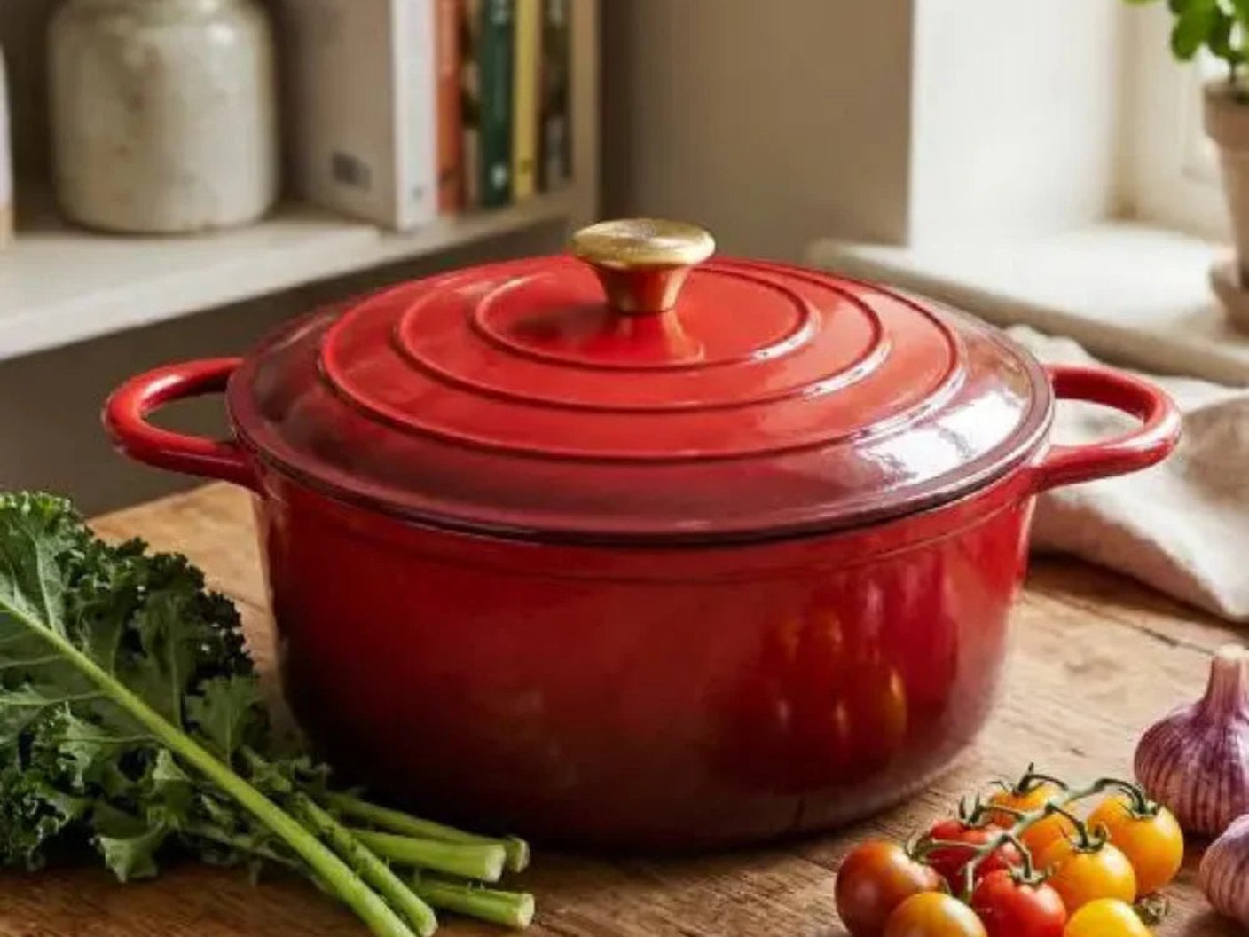 A red enamelled cast iron casserole on a timber counter with fresh vegetables in warm daylight