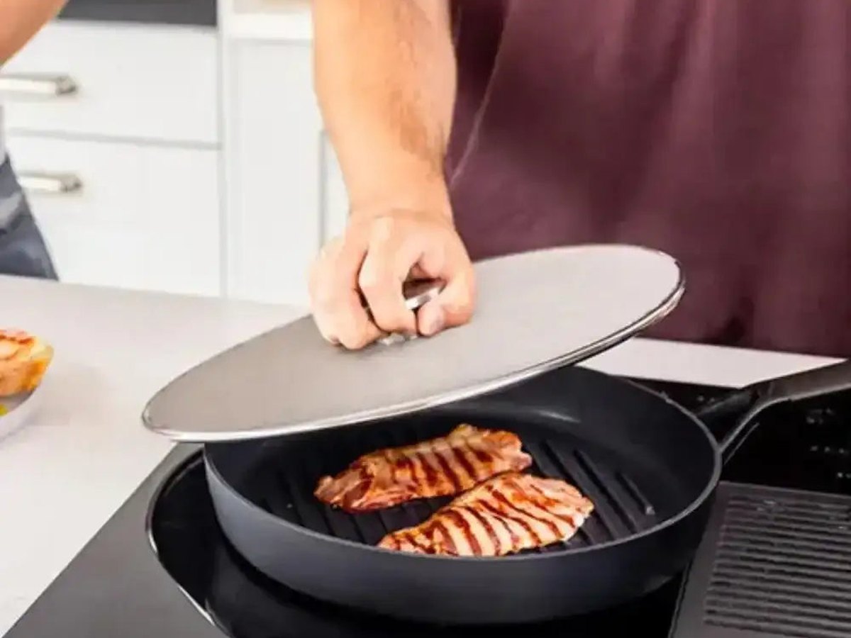 A hand placing a stainless steel splatter screen over a pan of salmon in a bright kitchen