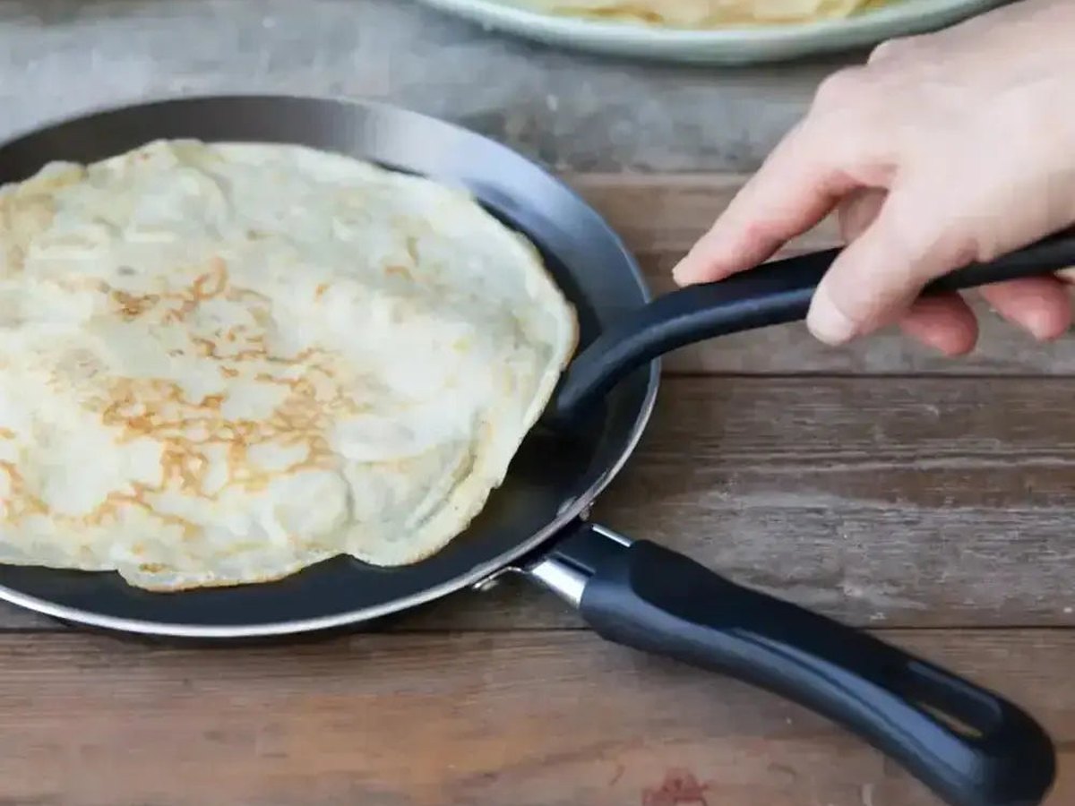 A hand tilting a non-stick pancake pan with a thin crepe, a stack of pancakes behind on a timber table