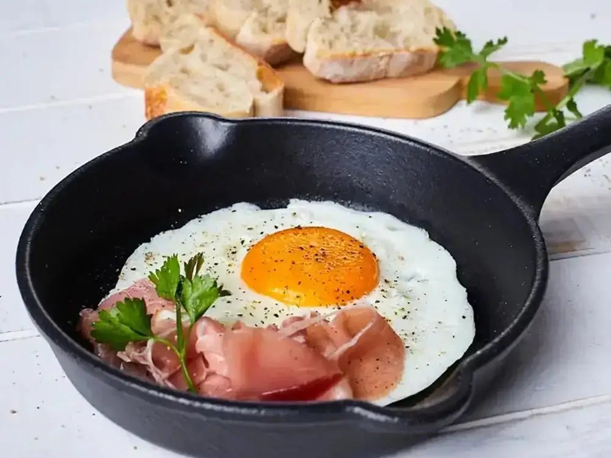 A fried egg and prosciutto cooking in a black enamelled cast-iron skillet, bread on a pale board in daylight