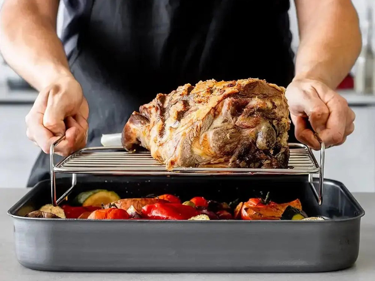 Hands lifting a roast joint on a stainless steel roasting rack from a tray of roasted vegetables