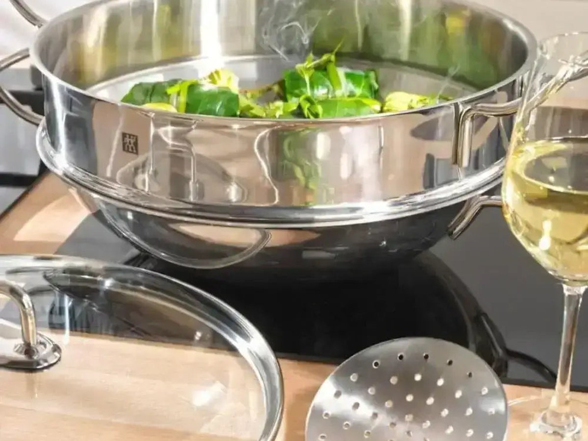 A polished stainless steel steamer filled with steaming green vegetables on an induction hob, its lid and skimmer resting on a timber board alongside