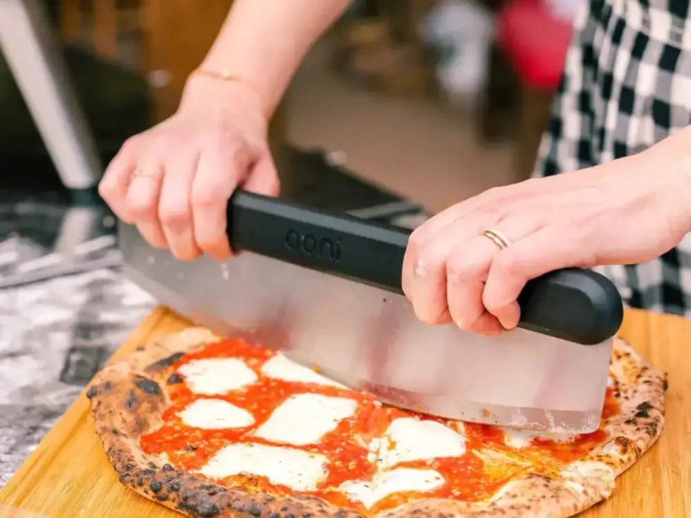 Hands cutting a wood-fired pizza with an Ooni rocker blade on a bamboo peel and serving board