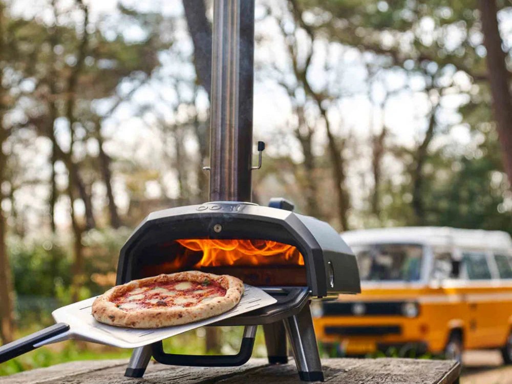 Ooni Karu 2 wood-fired pizza oven alight outdoors with a pizza on a peel in front of it