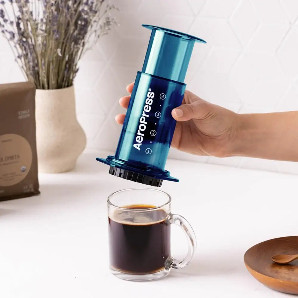 A person holding a blue Aeropress coffee maker over a glass of brewed coffee on a white surface.