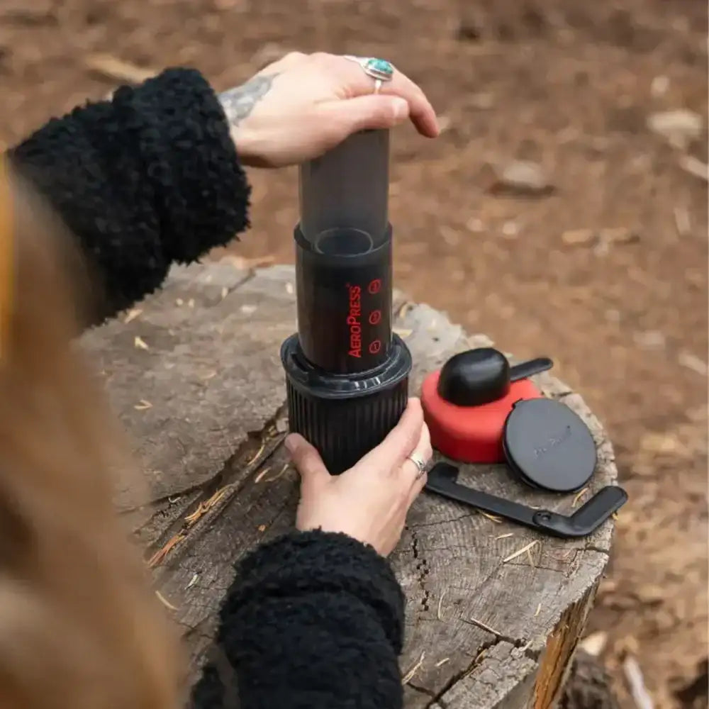 Person using a portable coffee maker on a wooden stump outdoors