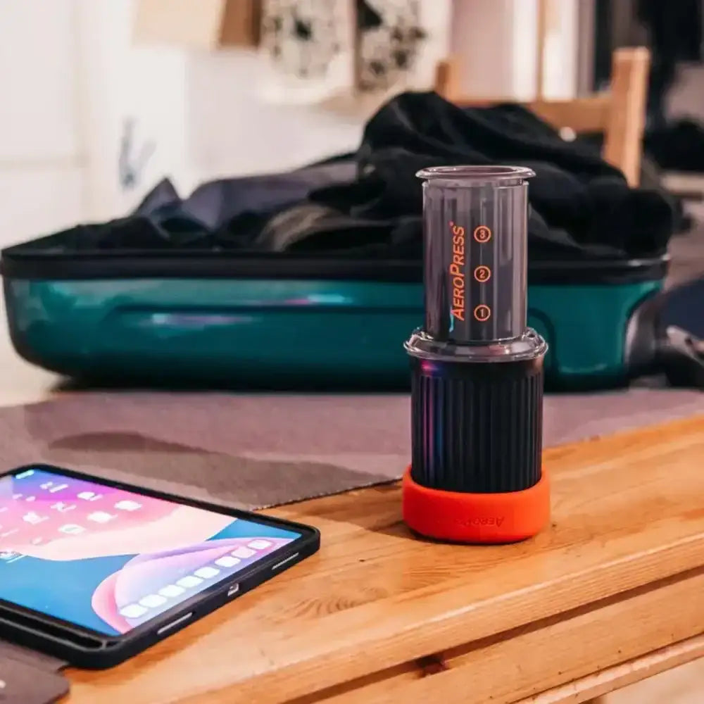 Aeropress coffee maker on a wooden surface with a phone and suitcase in the background