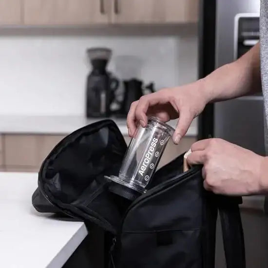 Person packing clear AeroPress coffee maker into black bag in modern kitchen