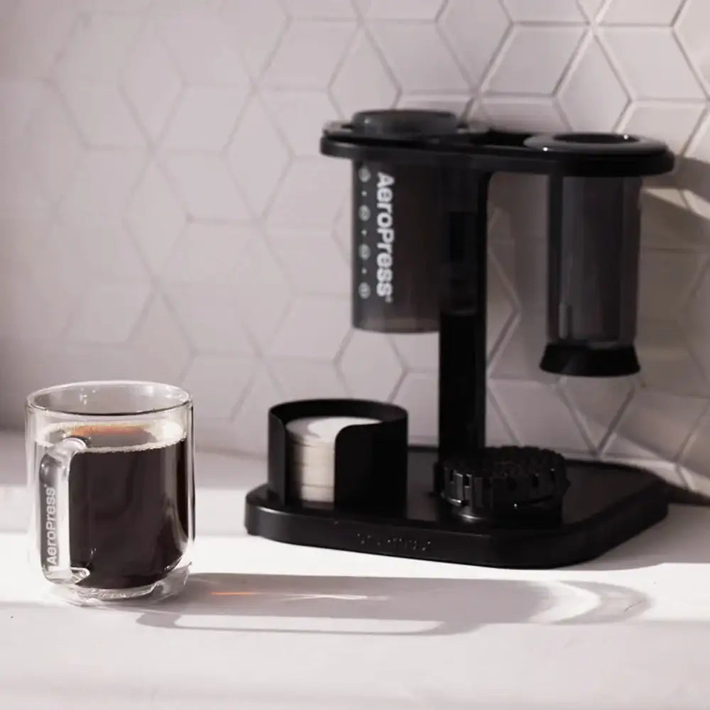 Aeropress coffee maker with a glass of coffee on a white surface.