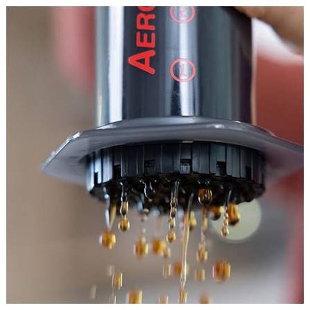 A close-up of a black AeroPress coffee maker with red branding, shown with liquid dripping from the plunger against a blurred background.