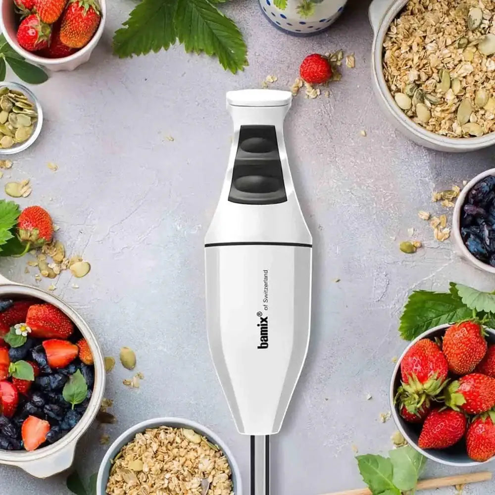 Handheld blender surrounded by bowls of fruits and granola on a gray surface