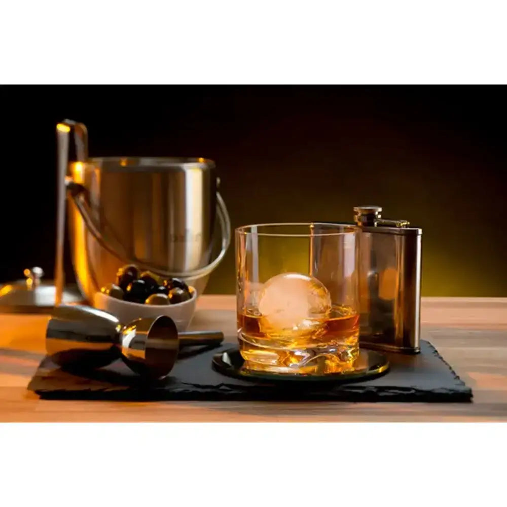 Glass of whiskey with a large ice cube on a wooden surface, surrounded by bar tools.