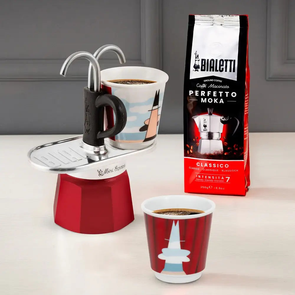 Red Bialetti Moka Express with two cups of coffee and a bag of Bialetti coffee beans on a gray surface.