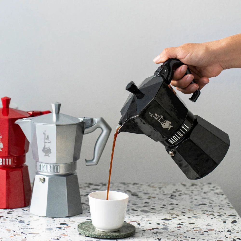 Person pouring coffee from a black Dallmayr coffee maker into a white cup on a speckled countertop.