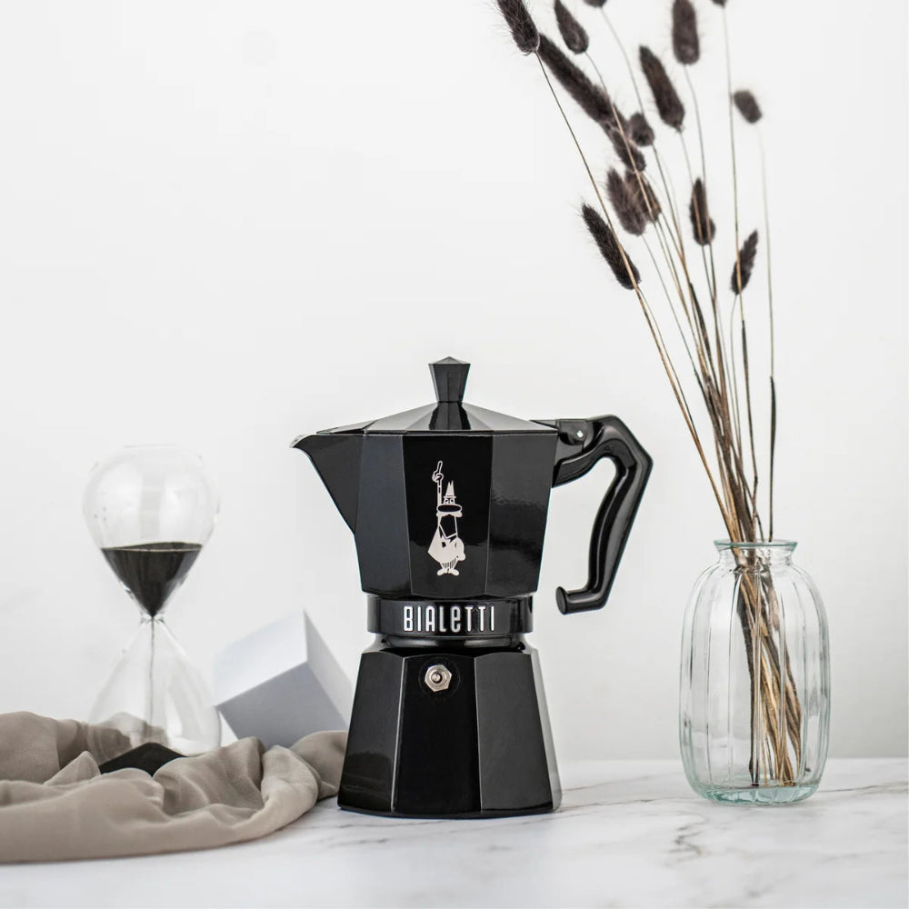 A black Bialetti moka pot coffee maker sits on a light marble surface, styled with a glass hourglass, a beige cloth, and a glass vase with dried stems against a white background.
