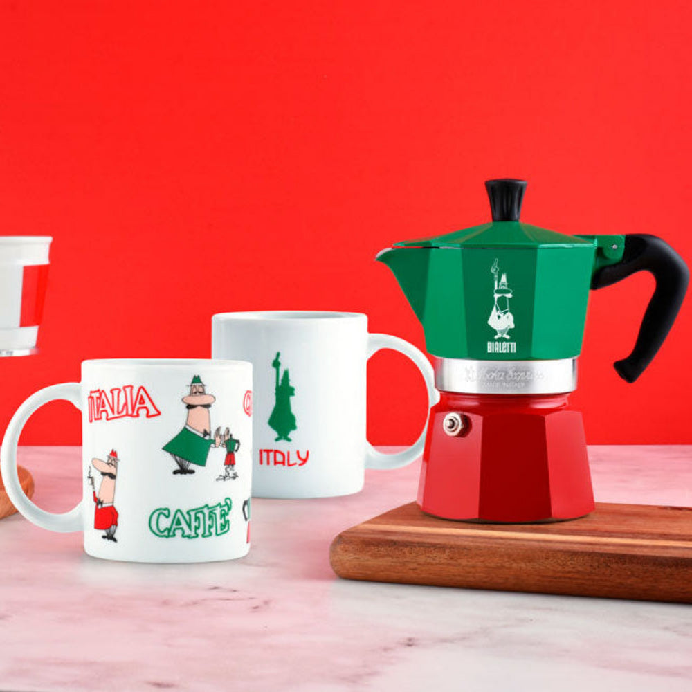 A red and green moka pot sits on a wooden board on a marble surface, with two white ceramic mugs featuring Italy-themed cartoon graphics in the background against a solid red wall.