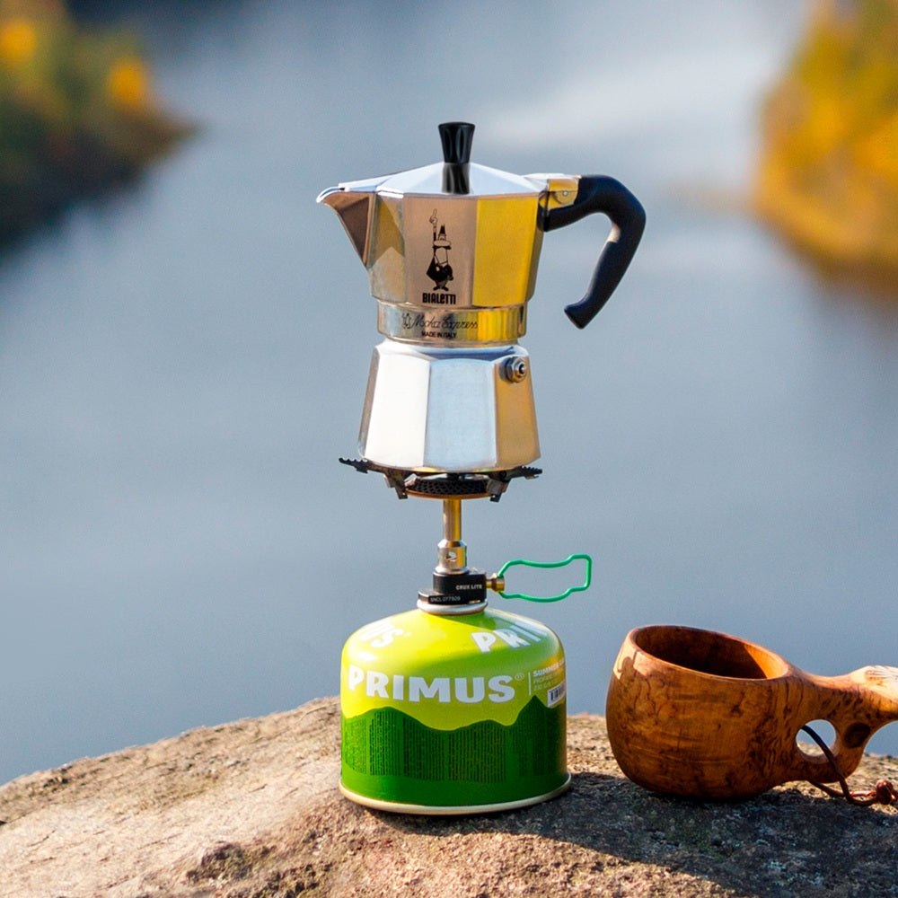 Portable coffee maker on a Primus stove with a wooden cup by a body of water.