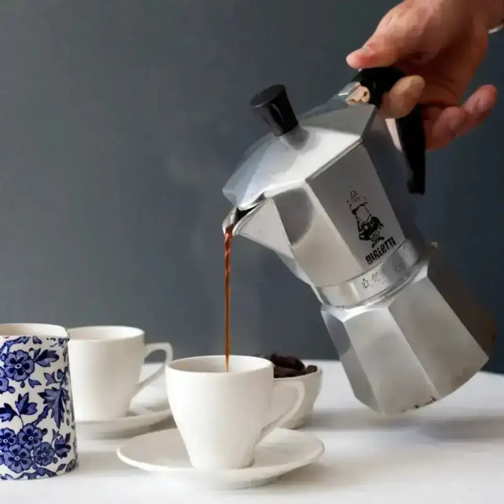 Stainless steel Moka pot pouring coffee into a white cup on a table with a dark background