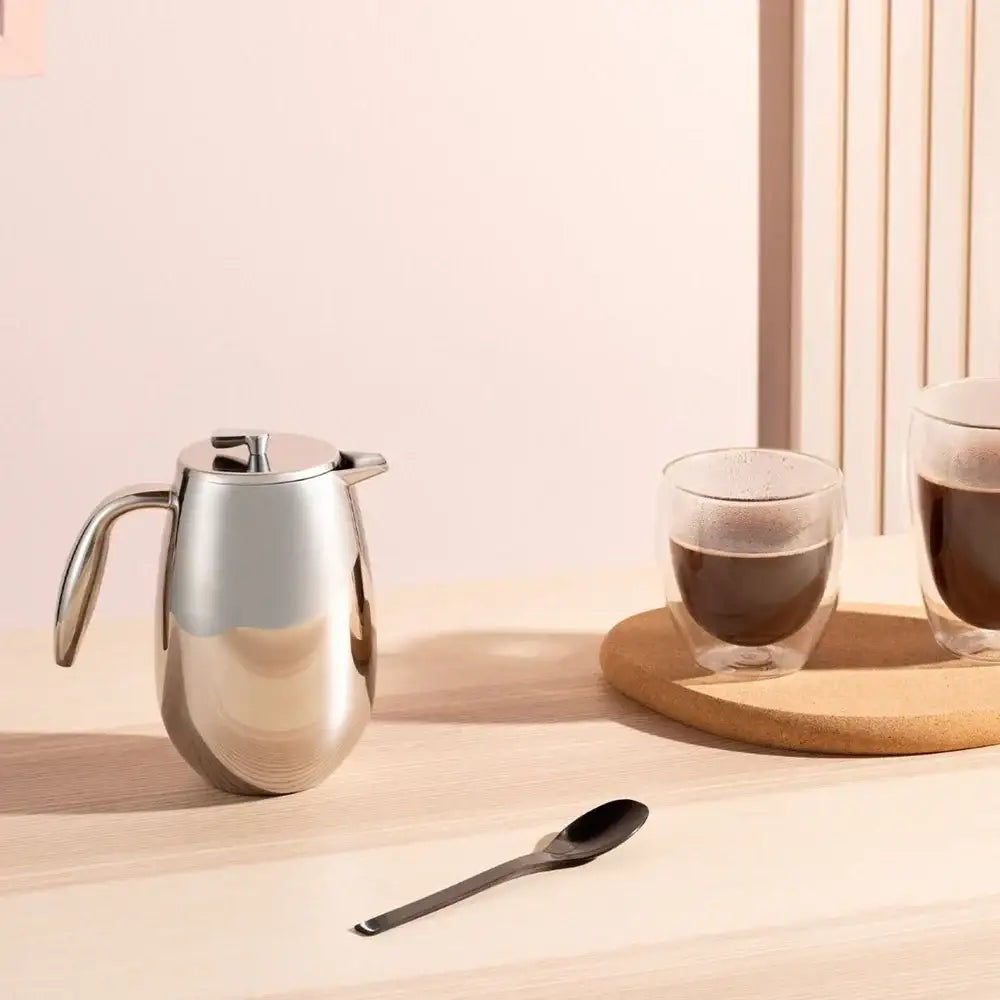 french press with a glass with coffee, and spoon on a wooden surface with a soft pink background