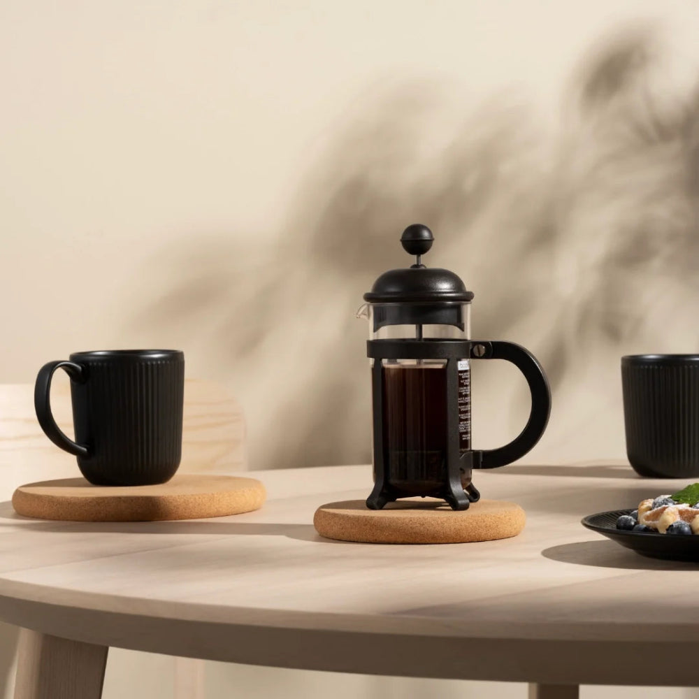 A black glass and metal French press sits on a round cork coaster on a light wood table, flanked by two black ceramic mugs on matching coasters. The scene is styled in a warm, minimalist setting with soft shadows cast on a beige wall.
