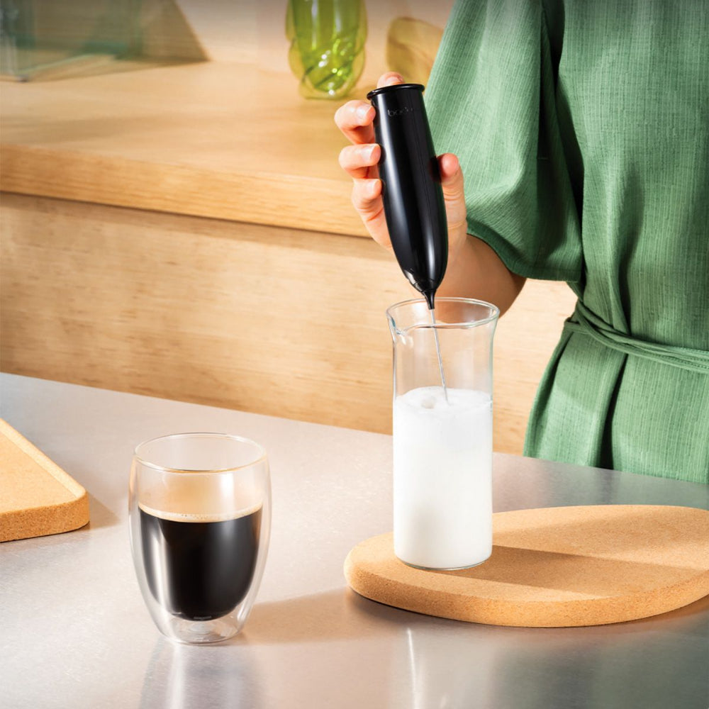 A person in a green dress uses a black handheld electric frother to mix milk in a clear glass pitcher on a wooden board, with a double-walled glass of dark coffee on a metallic countertop nearby.
