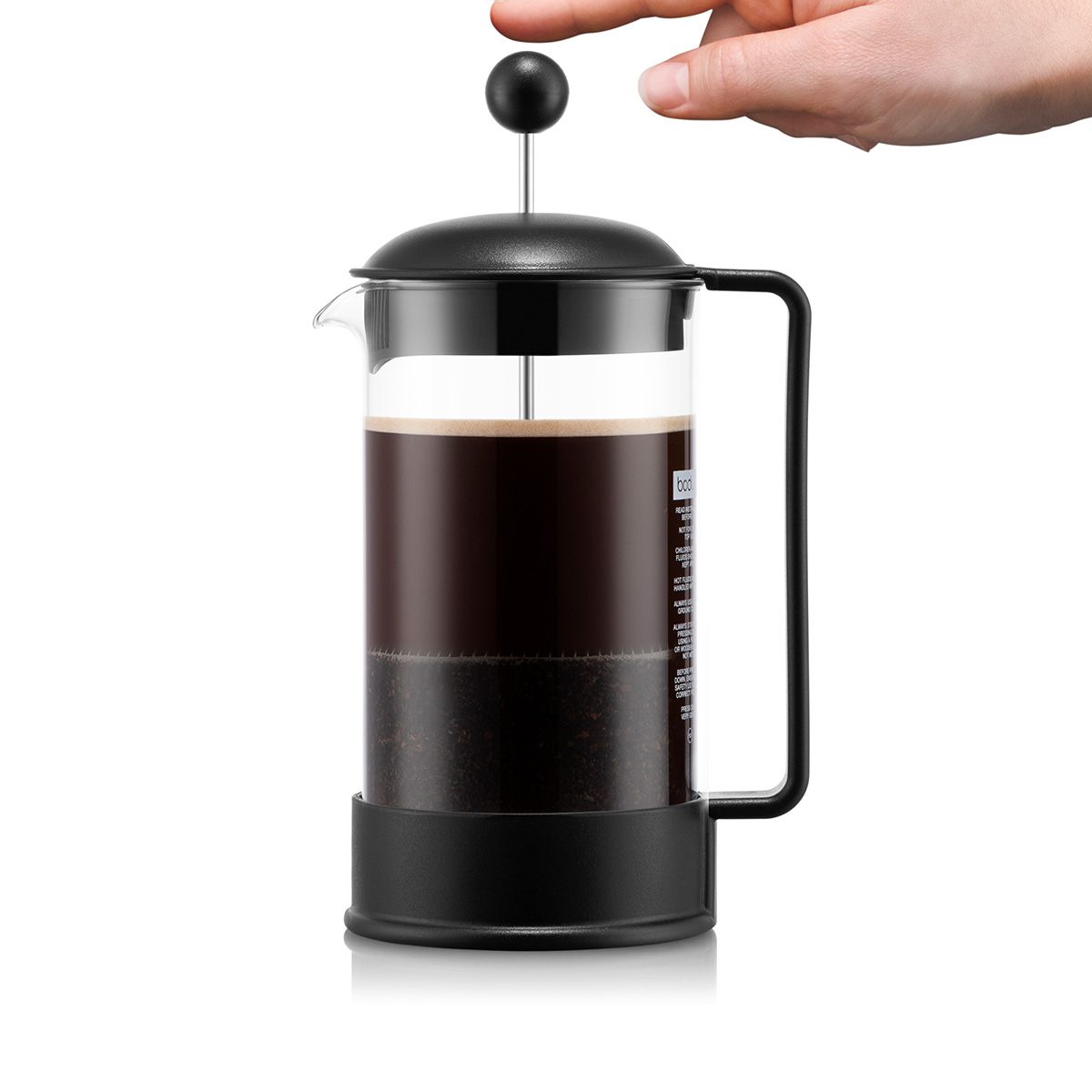 A classic French press coffee maker with a black metal body, handle, and lid, filled with dark coffee in a clear glass carafe, with a persons hand shown against a plain white background.