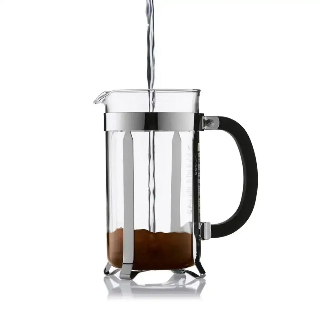 Clear glass French press with coffee grounds being filled with water on a white background