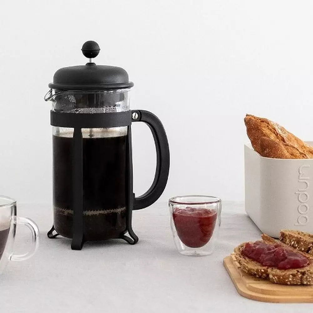 A black Bodum french press with a glass of brewed coffee, with a Bodum bread bin with bread on a bamboo lid on a white background