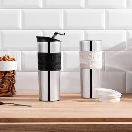Two silver travel mugs with black accents on a wooden surface against a white tiled wall.