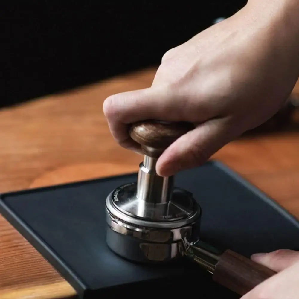 Hand using a coffee tamper on a black flat surface with a wooden table in the background