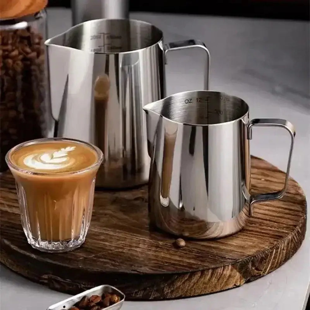 Two stainless steel milk pitchers on a wooden tray with a glass of coffee and coffee beans.
