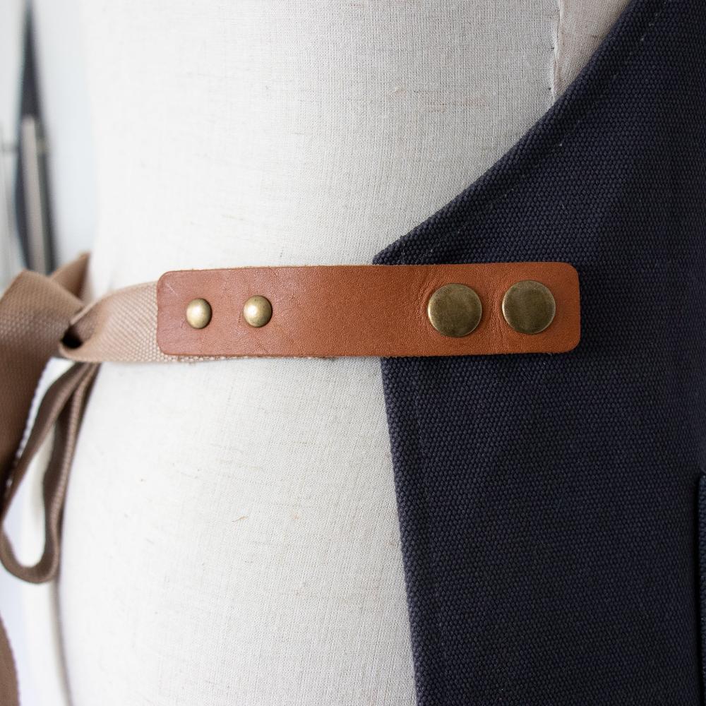 Navy apron with a brown leather strap and brass buttons on a mannequin.