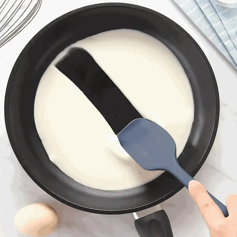 Person using a spatula to spread batter in a frying pan on a light surface.