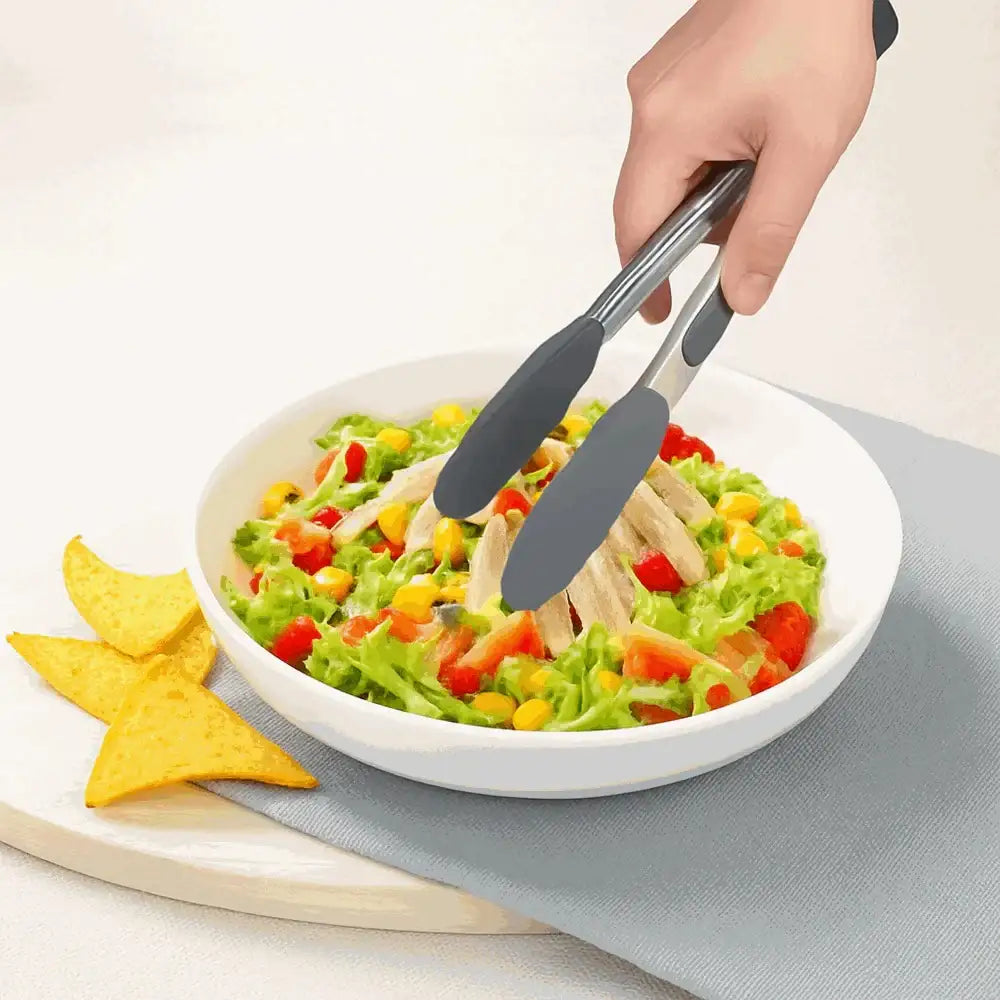 Hand using grey silicone kitchen tongs to serve salad from white bowl with tortilla chips