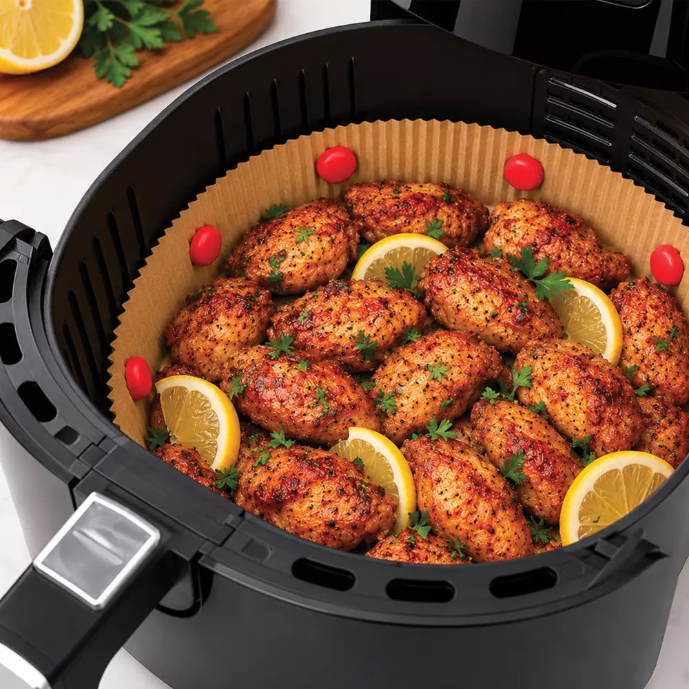 A black air fryer basket filled with seasoned chicken wings garnished with lemon slices, parsley, and cherry tomatoes, photographed on a light countertop.