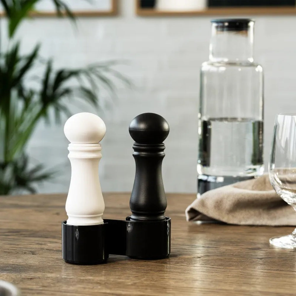 Black and white pepper grinders on a wooden table with a glass of water and napkin in the background.