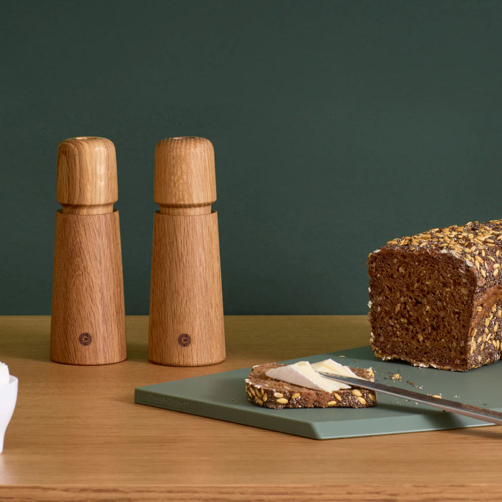 Salt and pepper grinders on a wooden surface with a loaf of bread on a cutting board