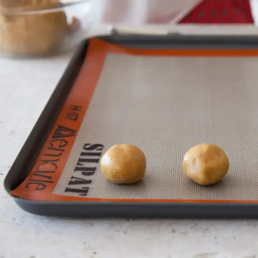 Two cookie dough balls on a Silpat baking mat with a glass bowl in the background.