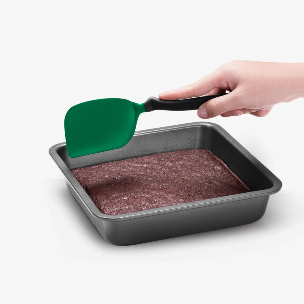 A hand holds a green silicone spatula with a black handle, lifting a square dark gray cake pan filled with brownies against a light gray background.