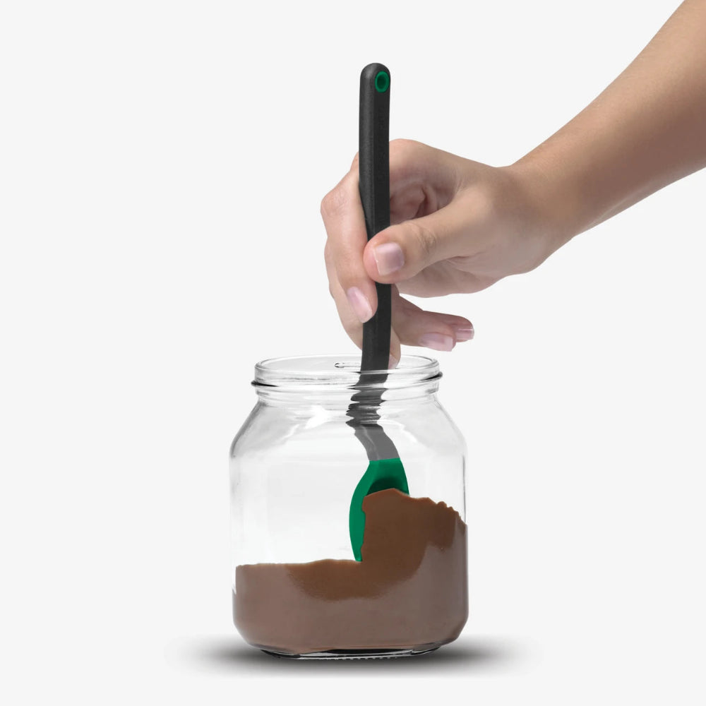 A hand holding a black-handled silicone spatula with a green spoon, stirring a brown cream inside a clear glass jar against a light gray background.