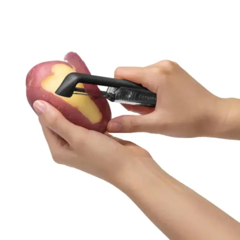 Person using a black potato peeler on a red potato against a white background