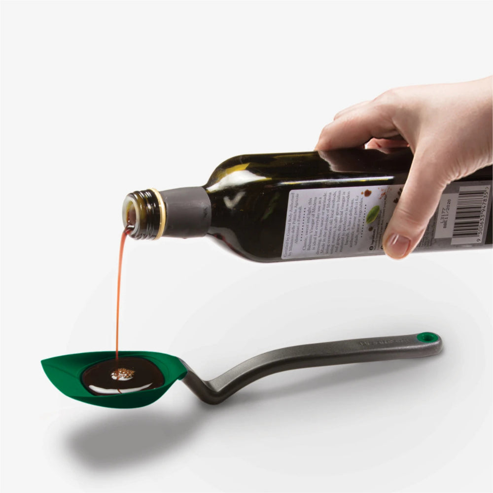 A hand holds a dark glass bottle and pours a stream of dark liquid into a green spoon resting on a gray spoon, against a clean light gray background.