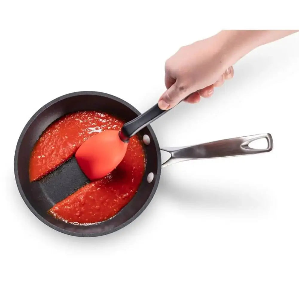 Person using a red spatula to stir tomato sauce in a black frying pan on a white background