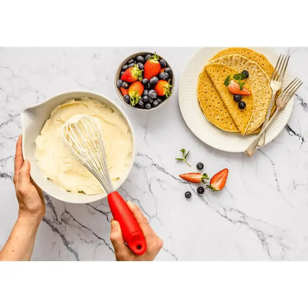 Person whisking batter with a red whisk, next to a plate of pancakes and a bowl of berries on a marble countertop.
