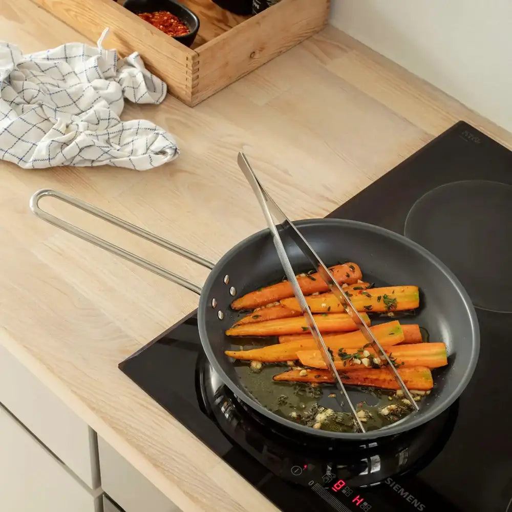 Cooking carrots in a pan on a stove with a wooden cutting board and towel in the background.